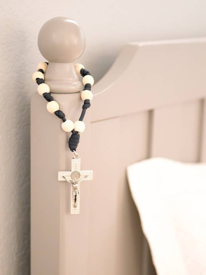 A door rosary with a silver-tone crucifix and St. Benedict Medal, featuring knotted beads in navy and gray colors, hanging on a bed post.