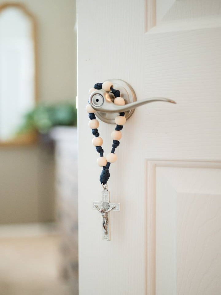 A door rosary with a silver-tone crucifix and St. Benedict Medal, featuring knotted beads in navy and gray colors, hanging on a door.