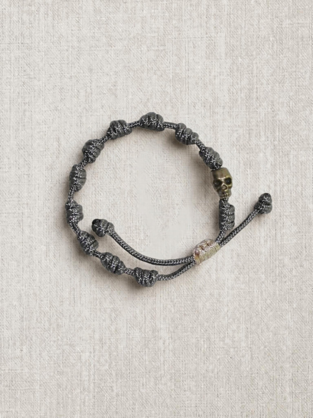 A dark gray knotted cord bracelet with a small metallic skull charm, displayed on a textured beige linen background.