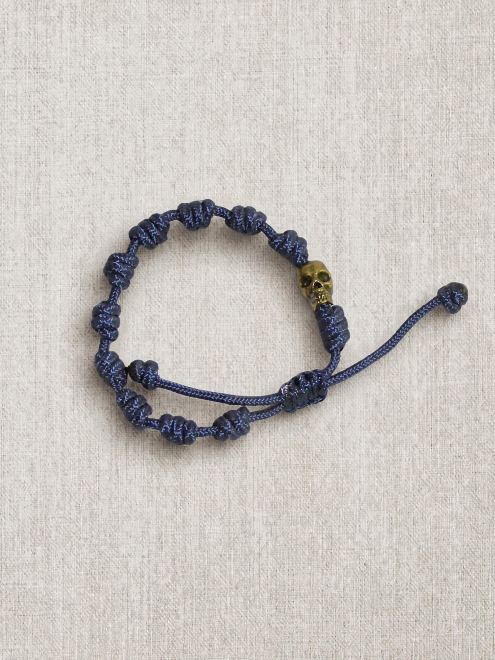 A dark blue knotted cord bracelet with a small gold skull accent bead, displayed on a textured beige linen background.