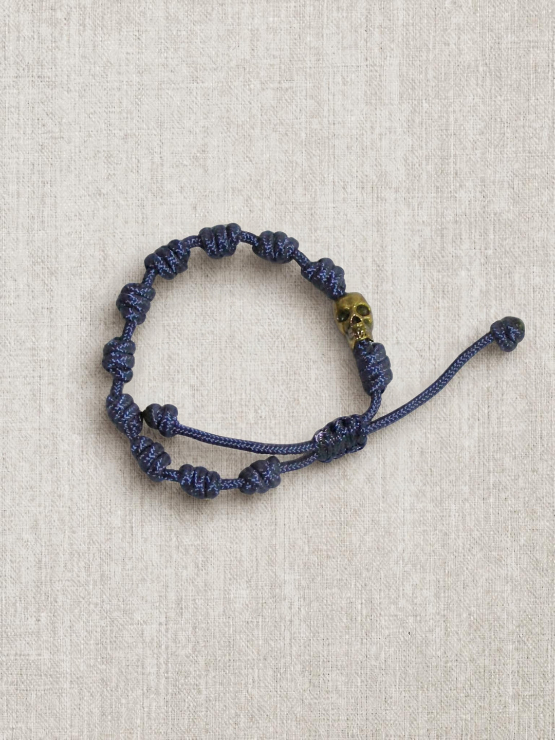 A dark blue knotted cord bracelet with a small gold skull accent bead, displayed on a textured beige linen background.