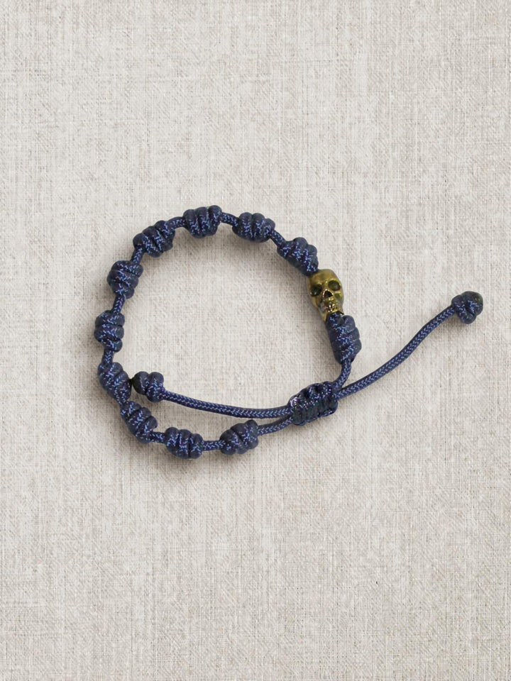 A dark blue knotted cord bracelet with a small gold skull accent bead, displayed on a textured beige linen background.