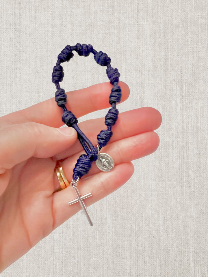A hand holding a dark purple knotted cord bracelet with a silver cross, a silver oval religious medallion, and a small gold ring, set against a light beige textured fabric background.
