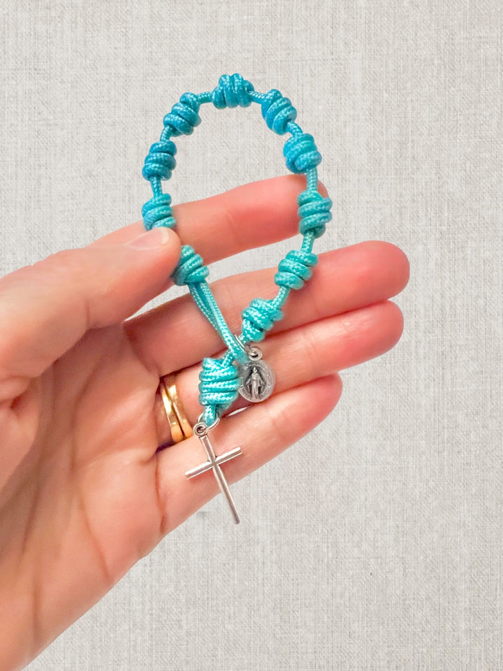A turquoise blue knotted cord bracelet with silver-tone religious charms, held in a person&