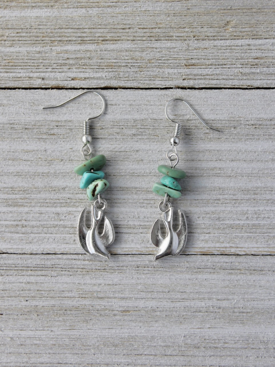 A pair of handmade silver dangle earrings featuring stacked natural turquoise stone chips and a shiny silver-tone dove charm representing the Holy Spirit, displayed on a rustic white wood background.